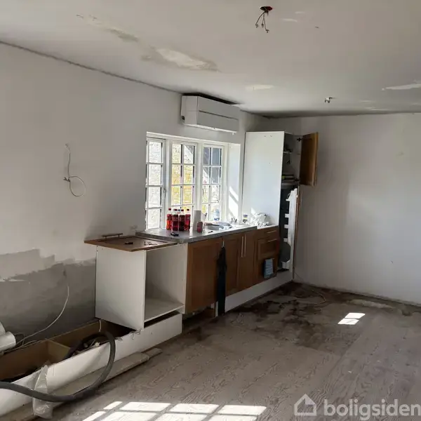Et ufuldstændigt køkken med manglende skabe og apparater står i et tomt rum. Vindue med persienner, rod på bordpladen samt en åben skabsdør giver indtryk af en renovering i gang.