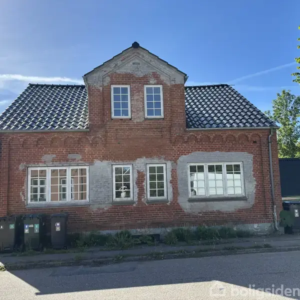 Rødstenshus med sort tegltag og hvide vinduer står i sollys ved en vej. Omkring huset er der grønt løv fra træer og buske samt skraldespande langs facaden.
