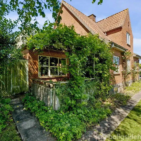 Et rødt murstenshus delvist dækket af grønne planter står i en forhave. En sti leder forbi huset til biler parkeret i baggrunden i et solrigt boligområde.