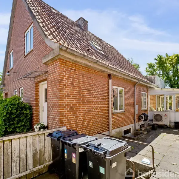 Et murstenshus står stille i en solrig baghave. Affaldscontainere er placeret foran, og en lille veranda er synlig til højre. Omkring er flisebelægning og grønne træer.