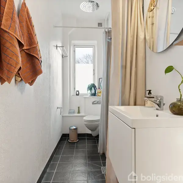 Badeværelse med hvidt vaskeskab, grøn plante i glasvase, bruseskærm og hvidt badeforhæng. Orange håndklæder hænger på væggen. Smalt vindue giver lys. Mørke gulvfliser og hvid væg.