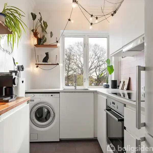 Køkken med vaskemaskine og ovn, træhylder med planter og figurer, vindue med udsigt til træer. Lyse vægge, træbord og hængende lamper skaber en hyggelig atmosfære.