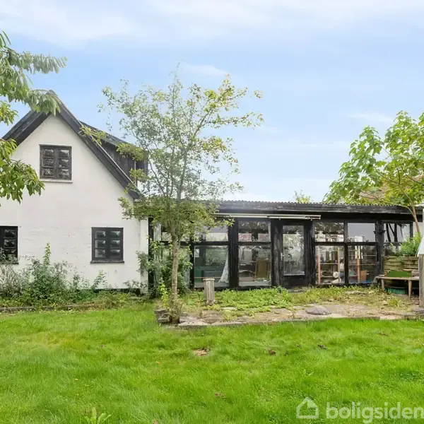 Hus med hvid facade og mørkt tag står stille i en grøn have med træer omkring. Store vinduer viser indvendige detaljer, og en lille terrasse er synlig foran huset.