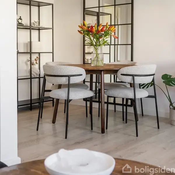 Rundt spisebord med fire stole omringet i et moderne rum. Glasvase med blomster står på bordet. Bagved ses en hylde med dekorative genstande og en grøn plante ved siden af.