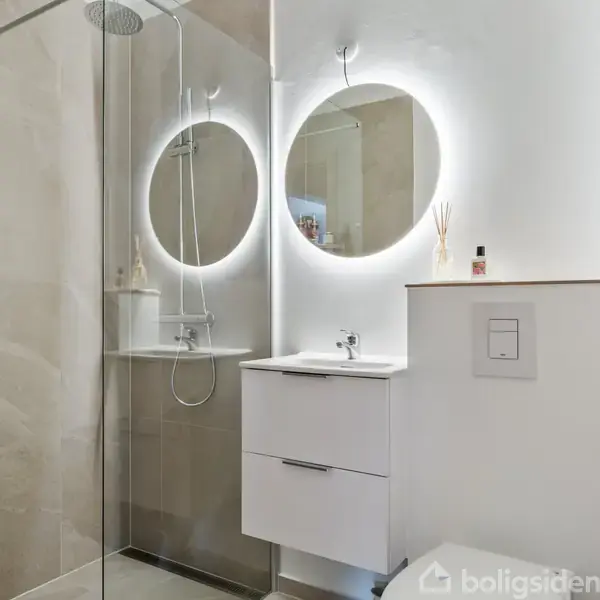 Et moderne badeværelse med en bruseniche til venstre. En rund lampe lyser et spejl op over en hvid vask. Toilettet er til højre i et stilrent rum.