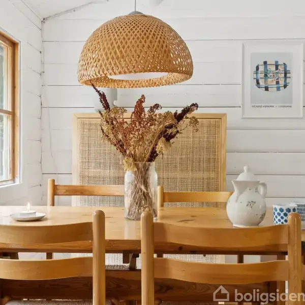 Et spisebord af træ med en vase med tørrede blomster står i et lyst rum med hvide trævægge og store vinduer. En flettet lampe hænger over bordet. På væggen er der et kunstværk med teksten "HOLMEGAARD VINTERBLOMST".