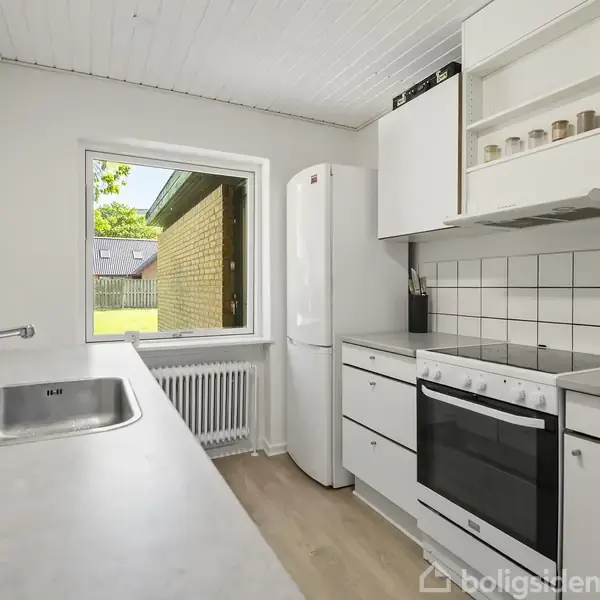 Et moderne køkken har en hvid bordplade med vask, et komfur og en høj køleskab. Det er omgivet af hvide skabe. Et vindue giver udsigt til en have.