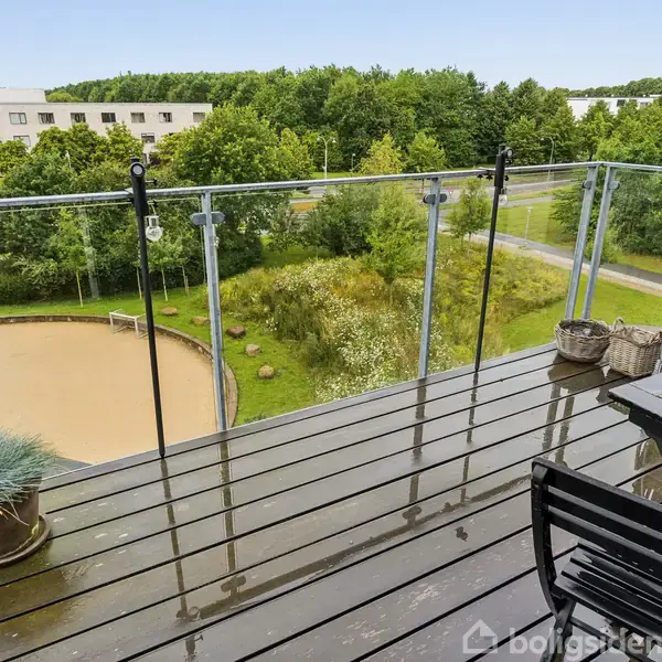 Balkon med sorte havemøbler står stille overfor en grøn park. Området inkluderer en sandjord sportsbane og flere træer i baggrunden.