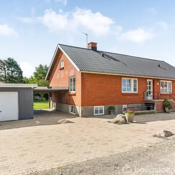 Et rødt murstenshus med sort tag står ved siden af en garage. Husets indgang har en trappe, og omgivelserne er en åben gårdsplads med brosten og spredte planter.