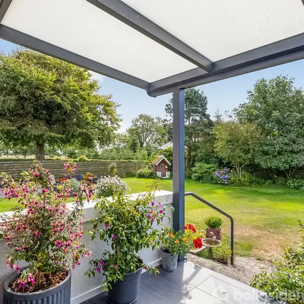 Terrassen har blomster i krukker, der står stille, omgivet af en grøn have med græs og store træer i baggrunden. En lille træbygning ses i haven.