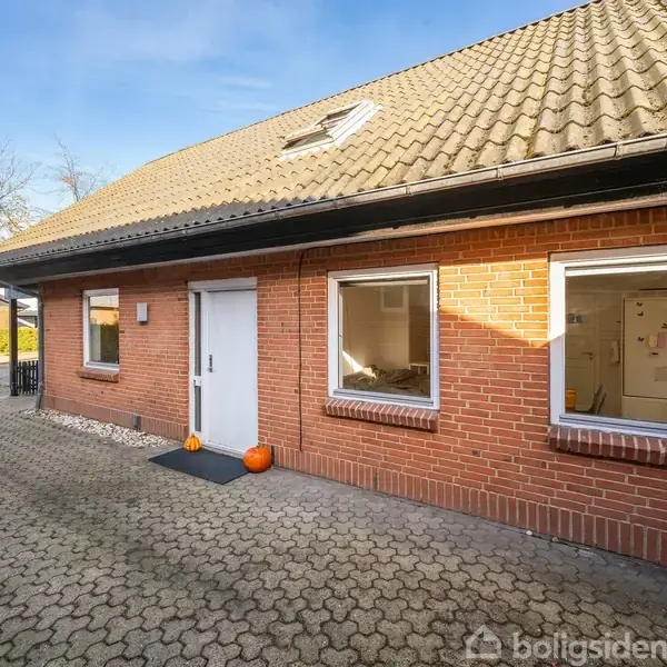Rødt murstenshus med skråt tag, står stille. To græskar ved hoveddøren. Omgivet af flisebelagt gårdsplads, ses en garage til venstre og træ bagved. Ingen tekst.