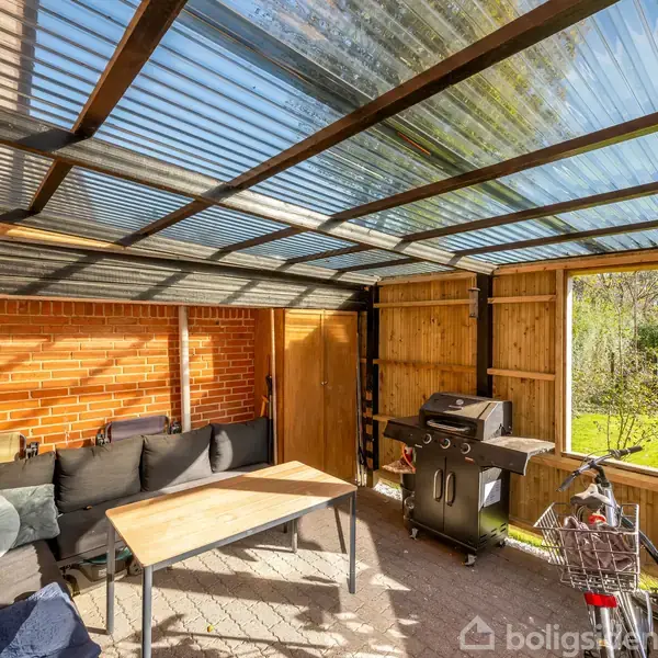 Terrasse med tag, sofa og bord; en grill står ved væggen. Cykkel og kurv ses i forgrunden. Sollys oplyser stedet, og have ses udenfor.