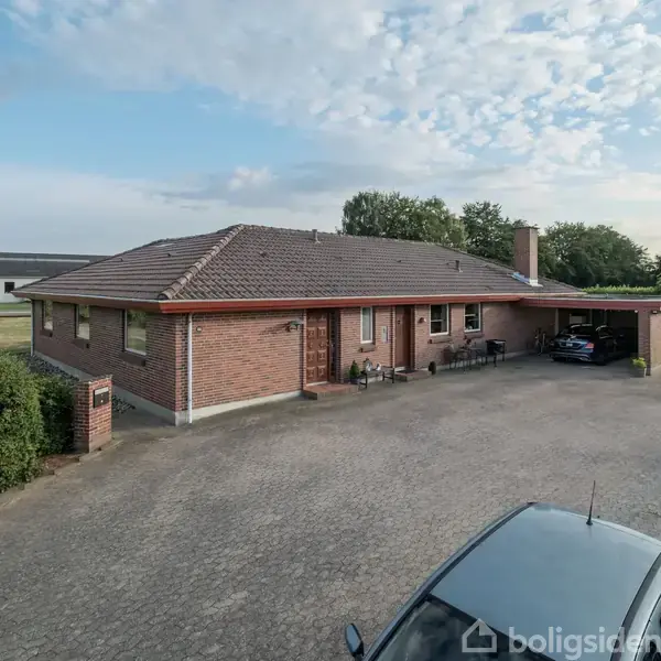 En rød murstensvilla med skråt tag, parkeret under en carport. Foran huset er der en stor asfalteret gårdsplads med en bil. Baggrunden viser grønne buske og træer.