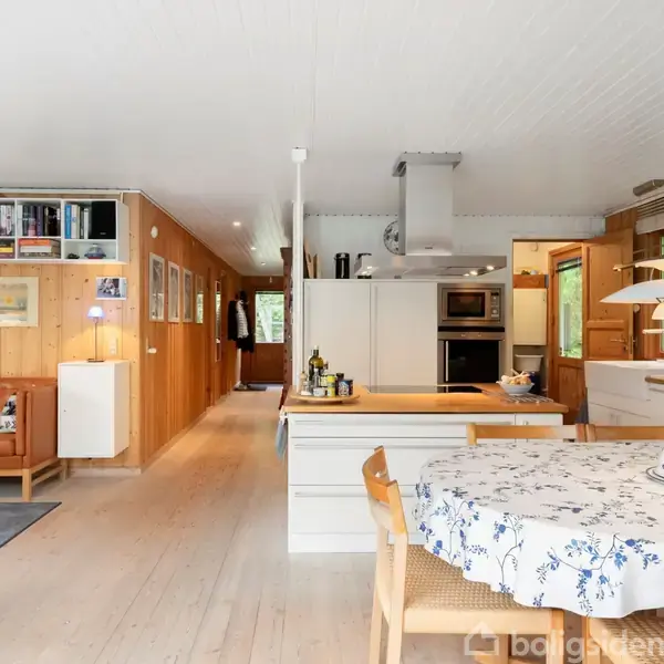 Køkken-alrum med spisebord dækket af hvidt og blåt mønstret dug, omgivet af træstole. Åben stue til venstre med sofa og reoler, og moderne køkken til højre.