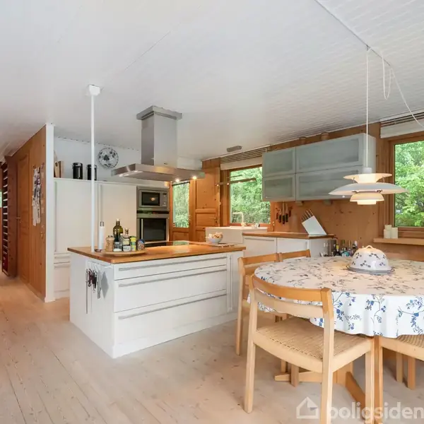 Køkken med hvid køkkenø og komfur, omgivet af træpaneler og store vinduer, der giver udsyn til grøn natur. Rundt spisebord med blomsterdug og fire stole.