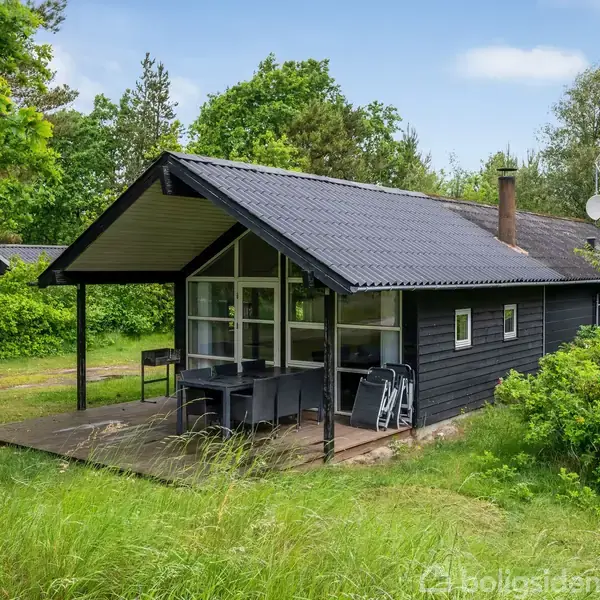 Sort træhus står stille på en grøn græsplæne, omgivet af træer. Der er en overdækket terrasse med bord og stole. Skorsten peger op fra tagets centrum.