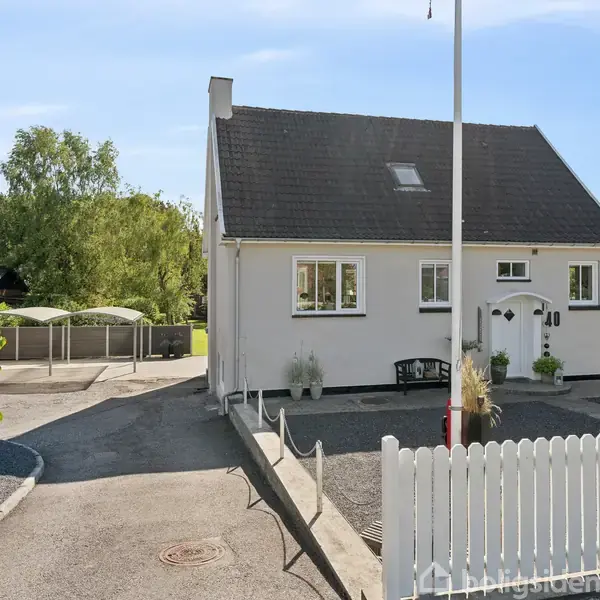 Et hvidt enfamiliehus med mørkt tag står bag et hvidt stakithegn. Huset har vinduer og en dør med dekorative blomster. Omkringliggende træer ses på en solrig dag.