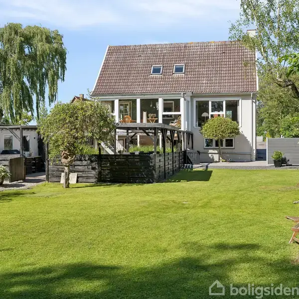 Et stort græsareal er omgivet af træer og buske, foran et hvidt hus med rødt tag. Der er træstole og en pergola i haven.