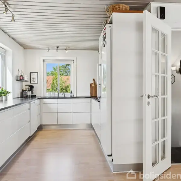 Køkken med hvide skabe og sorte bordplader strækker sig langs væggen. Et åbent dørparti fører til en gang med et rundt spejl. Et vindue giver udsigt til træer udenfor.