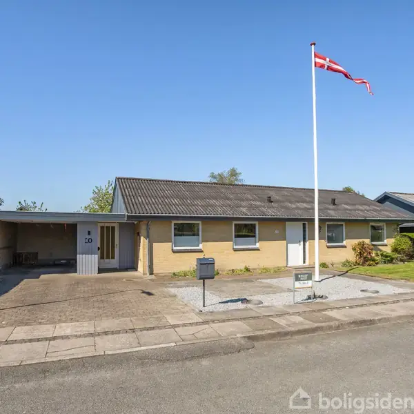 En enkelt seng står mod en hvid væg, ved siden af et skrivebord og stol. Rummet er lyst op af en loftslampe, og der er et vindue til venstre.