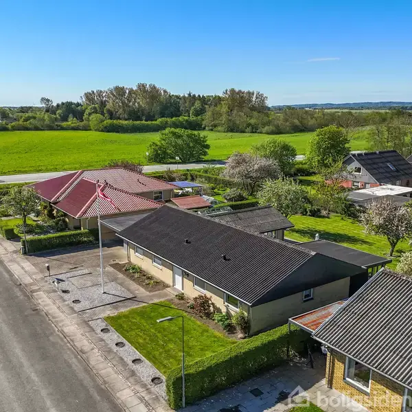 Et parcelhus med sort tag står midt i et villakvarter. Græsplæne og kantet hæk omkranser huset. Baggrunden viser grønne marker og træer under en klar blå himmel.