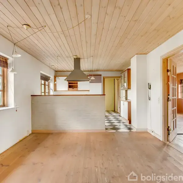 Køkkenalrum med træloft, emhætte over halvåben murstensvæg, omkranset af vinduer. Køkkenet har skakternet gulv, og en dør fører til et andet rum med trægulv og store vinduer.