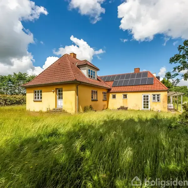 Gult hus med rødt tag og solpaneler står på en grøn græsplæne omgivet af tætte buskadser under en blå himmel med hvide skyer.