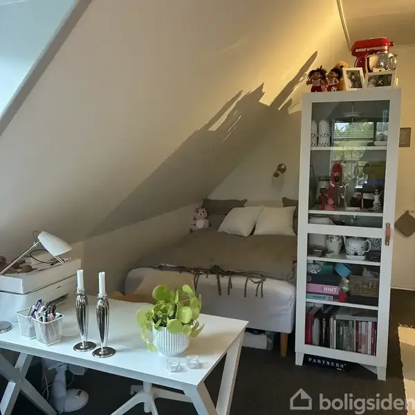 En seng med gråt sengetøj står i hjørnet af et værelse med skrå vægge. Ved siden af er en skabsreol fyldt med bøger og pynt. Et bord med planter og lysestager står i forgrunden.