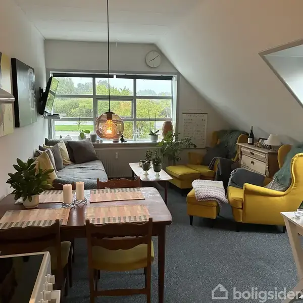 Et hyggeligt stueområde med en grå sofa og to gule lænestole ved et stort vindue. Et træbord med planter og stearinlys står ved siden af, og der er en hængende lampe.