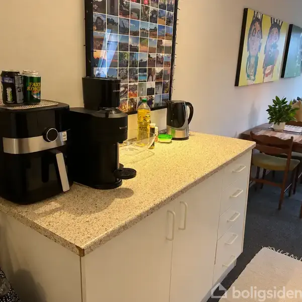 En køkkenbordplade med en airfryer og kaffemaskine placeret i et køkken-alrum. Baggrunden viser en spisestue med træmøbler og dekorationer på væggene.