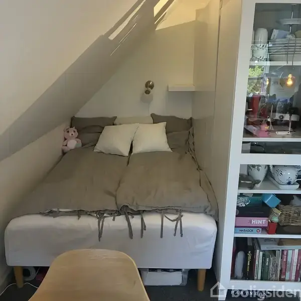 En seng med gråt sengetøj, placeret under en skråvæg. En lyserød bamse ligger ved hovedenden. Til højre er der en hylde og opbevaringsenhed med spil og bøger.