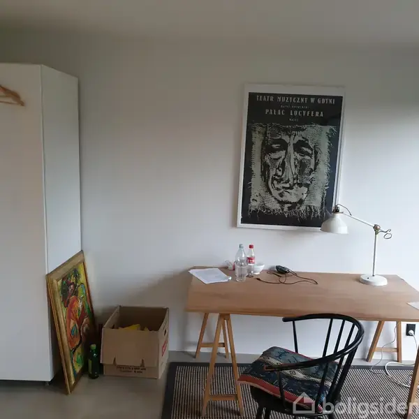 Et skrivebord står foran en væg med en lampe og papirer, flankeret af en stol. Over bordet hænger et portrætmaleri med teksten "Peter Brandes og Peter Laugesen". En hvid garderobe står til venstre.