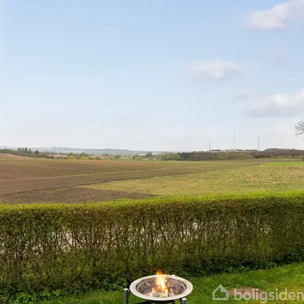 En udendørs ildsted brænder stille foran en lav hæk i et åbent landskab, hvor marker og træer strækker sig mod horisonten under en blå himmel med lette skyer.