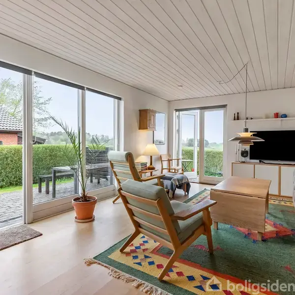 Stue indeholder sofa, lænestol og sofabord, med udsigt til have gennem store vinduer. Udenfor står havemøbler på terrasse. Rum dekoreret med tæppe og hylde med TV.