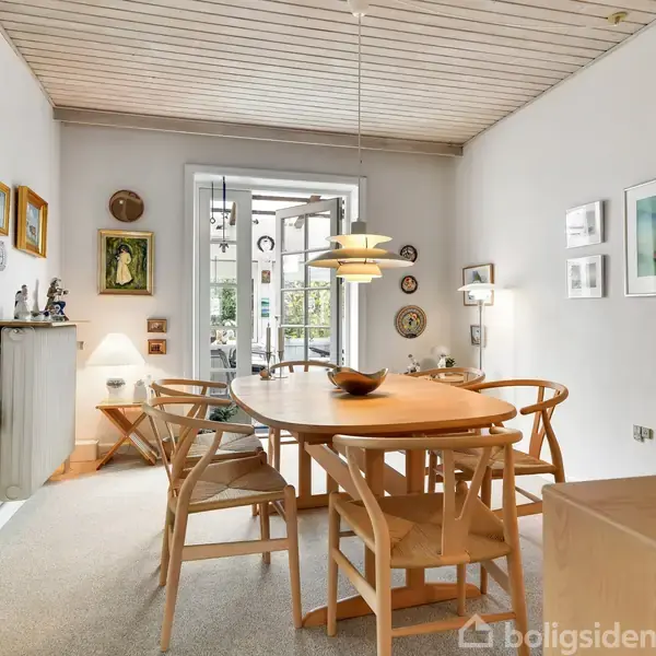 Rundt spisebord med seks stole står i et lyst rum med kunst på væggene. Lampe hænger over bordet, og en glasdør fører til en terrasse med grønne planter udenfor.