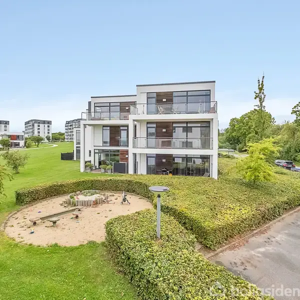 En treetagers moderne boligbygning står omgivet af en velholdt græsplæne og en sandkasselejeplads. I baggrunden er der flere lignende bygninger og grønne områder.