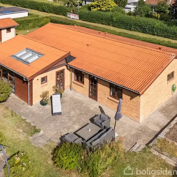 Huset er en enkeltplans murstensbygning med rødt tegltag og et tagvindue. Der er en terrasse med havemøbler og parasol. Haven er velplejet med buske og græsplæne.