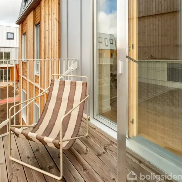 En stribet stol står på en lille balkon med trægulv. Omkring er moderne træbeklædte bygninger med store vinduer i en urban boligudvikling.