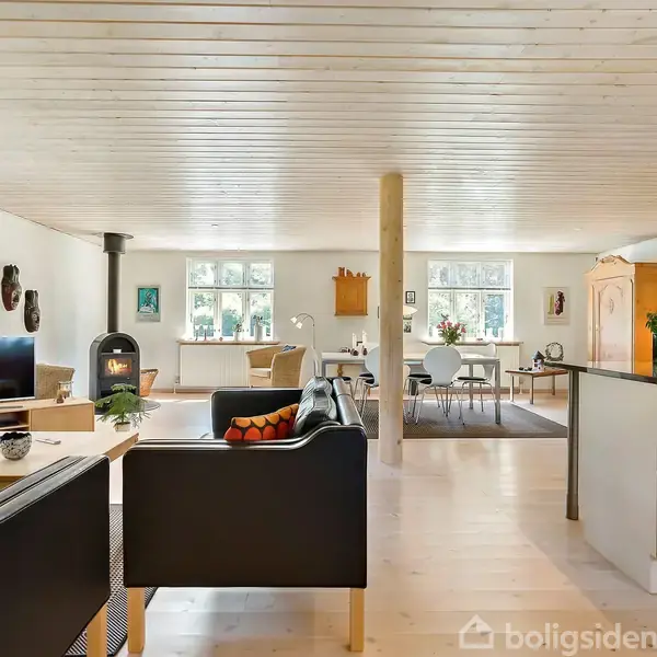 Stue med sorte sofaer placeret omkring et sofabord; pejs i baggrunden ved siden af et TV. Til højre ses et lyst køkken. Store vinduer giver naturligt lys.