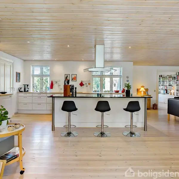 Et køkken har en central ø med tre sorte barstole. Køkkenet er lyst med hvidt træloft og store vinduer, omgivet af åbent opholdsrum med sofa og bogreol.