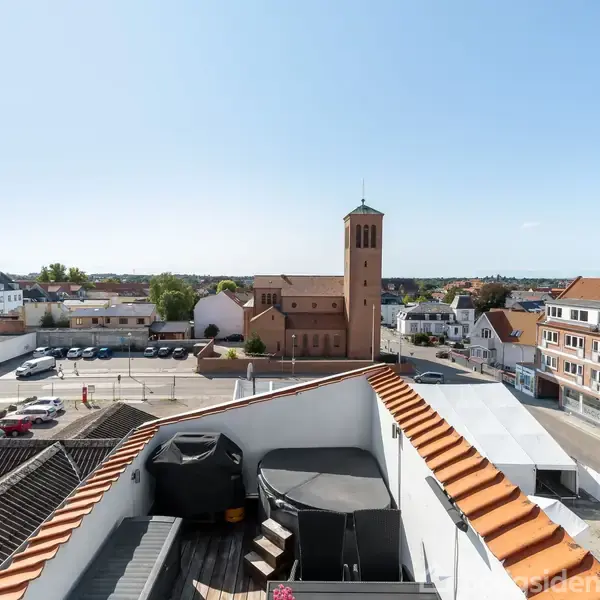 Tagterrasse med sorte møbler vender ud mod en byudsigt. Kirketårn og bygninger ses i baggrunden under en klar blå himmel.