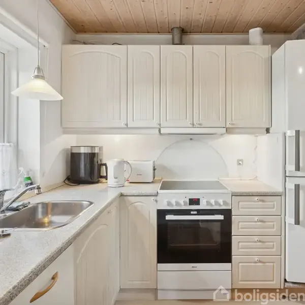 Et lyst køkken med hvide skabe og træloft. En ovn er centralt placeret mellem skabe, og der er en stor hvid køleskab ved siden af. En vask er ved et vindue.