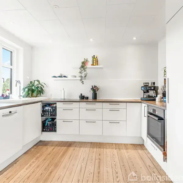 Et lyst køkken med hvide skabe og træbordplader. En kaffemaskine står på bordet ved siden af ovnen. Store vinduer til venstre bringer lys ind i rummet.