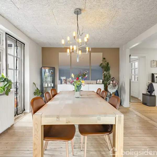 Et lyst spisestue bord omgivet af seks brune stole står i et rum med trægulv og moderne lysekrone. Rummet har store vinduer og en åbning til køkkenet. Der er planter ved vinduet og et stort maleri på væggen.