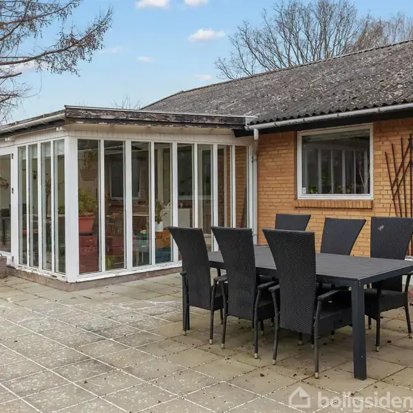 En sort spisebord med seks stole står på en flisebelagt terrasse foran et orangeri og et gult murstenshus. I baggrunden ses et træ og klar himmel.