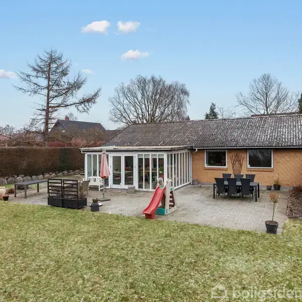 En enkelthus med en græsplæne og terrasse, hvor der står bord og stole. En rutsjebane fører ned til haven. Omgivet af hække og træer i et roligt villakvarter.
