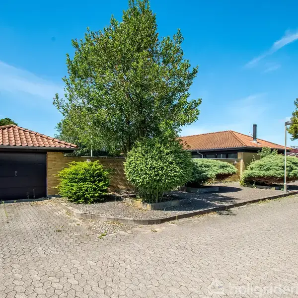 Et gult murstenshus med rødt tegltag ligger ved siden af en garage, omgivet af grønne buske og træer. Solrig dag med blå himmel ses i baggrunden.
