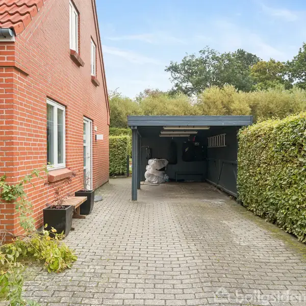 En murstensbygning står stillet op ad en brostensbelagt indkørsel. En carport ved siden af huset beskytter nogle genstande. Omgivet af en klippet hæk og træer i baggrunden.
