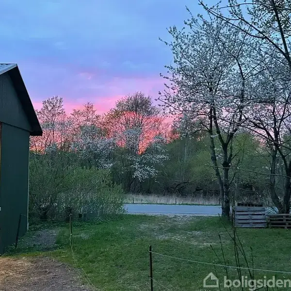En lade står åben mod venstre, omkranset af grønne træer, mens en vej er synlig i baggrunden under en rosa og blå solnedgang.