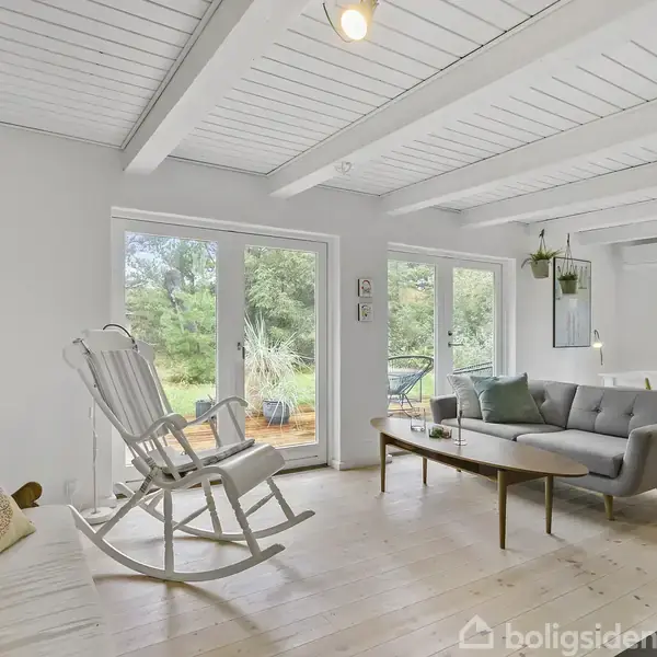 Gyngestol står i en lys, moderne stue med store vinduer, der giver udsigt til en terrasse og grønne omgivelser. Sofa, sofabord og spisebord ses i baggrunden.